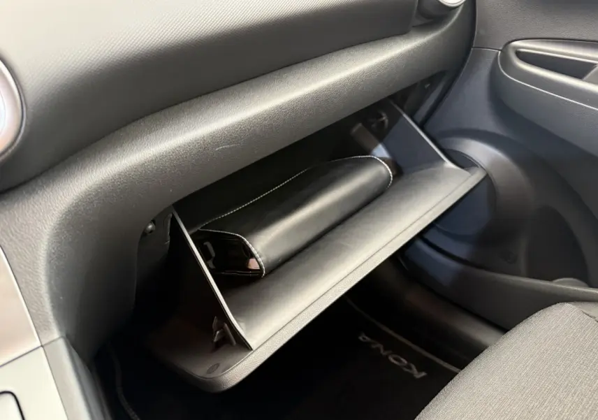 Intérieur du Hyundai Kona électrique 2022, vue sur la boîte à gants ouverte avec pochette noire et tapis de sol marqué Kona.