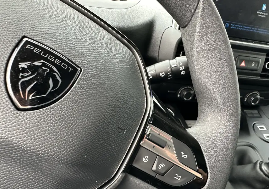 Gros plan sur le volant en cuir noir du Peugeot Partner Fourgon 2024 avec commandes intégrées et logo lion argenté.