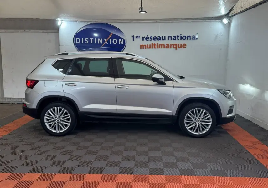 Profil côté droit d'un SUV SEAT Ateca gris clair 2019 avec jantes alliage et barres de toit chromées en intérieur showroom.
