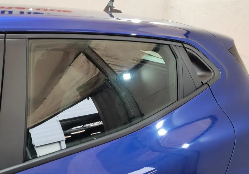 Détail de la vitre arrière côté gauche d'une Renault Clio bleu 2025 avec carrosserie lisse et reflet d'atelier visible.