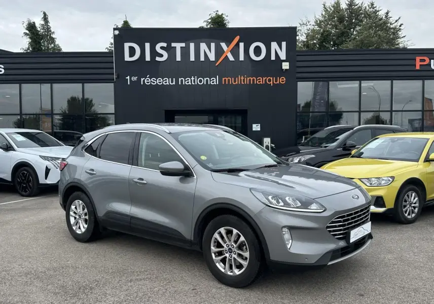 Ford Kuga gris solaire vu en 3/4 avant droit avec barres de toit aluminium sur parking devant un bâtiment Distinxion.