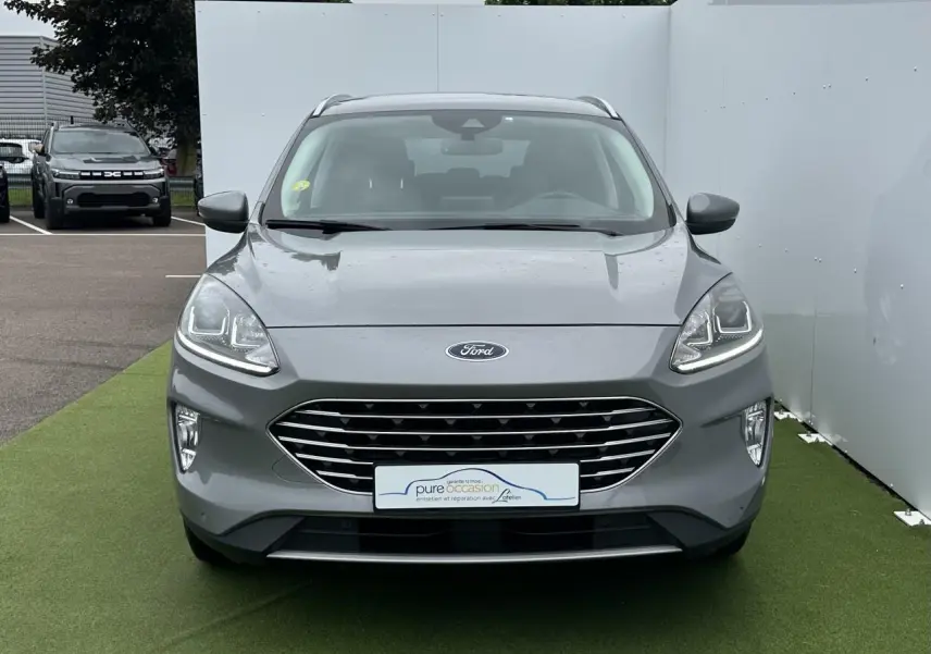 Vue de face d'un Ford Kuga gris solaire avec calandre chromée et feux LED allumés sur un sol vert.