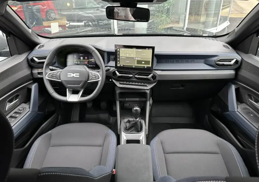Vue intérieure frontale du tableau de bord noir du Dacia Duster 2025 avec écran tactile et boîte manuelle.