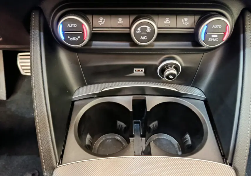 Gros plan sur la console centrale noire de l'Alfa Romeo Stelvio 2024 avec commandes climatisation et prises USB et 12V.