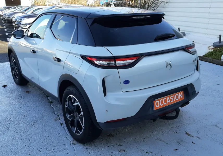 Vue 3/4 arrière droite d'une DS3 Crossback blanc perle avec toit noir, feux LED et attelage visible sur parking.