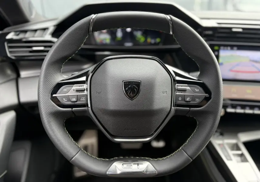 Volant cuir noir Peugeot 308 GT avec logo lion et badge GT, tableau de bord numérique et écran de caméra de recul visibles.