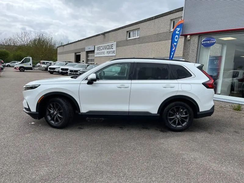 Vue de profil côté gauche d'un Honda CR-V blanc platine nacré avec jantes alliage noires, stationné devant un garage.