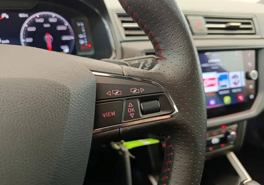 Gros plan sur le volant cuir noir avec surpiqûres rouges du SEAT Arona 2021, tableau de bord et écran tactile en arrière-plan.