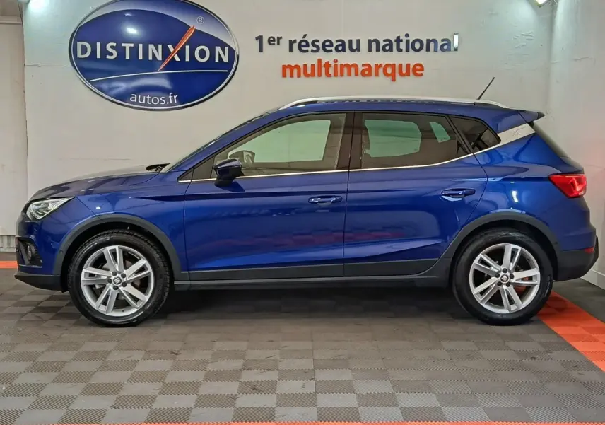 Profil côté gauche d'un SEAT Arona bleu 2021 avec jantes alliage et barres de toit chromées en intérieur showroom.