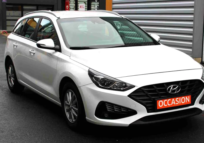 Vue 3/4 avant d'une Hyundai i30 SW blanche polaire avec calandre noire et plaque "OCCASION" rouge visible.