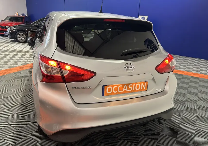 Vue 3/4 arrière d'une Nissan Pulsar gris métallisé avec feux arrière allumés et plaque "OCCASION" en intérieur showroom.