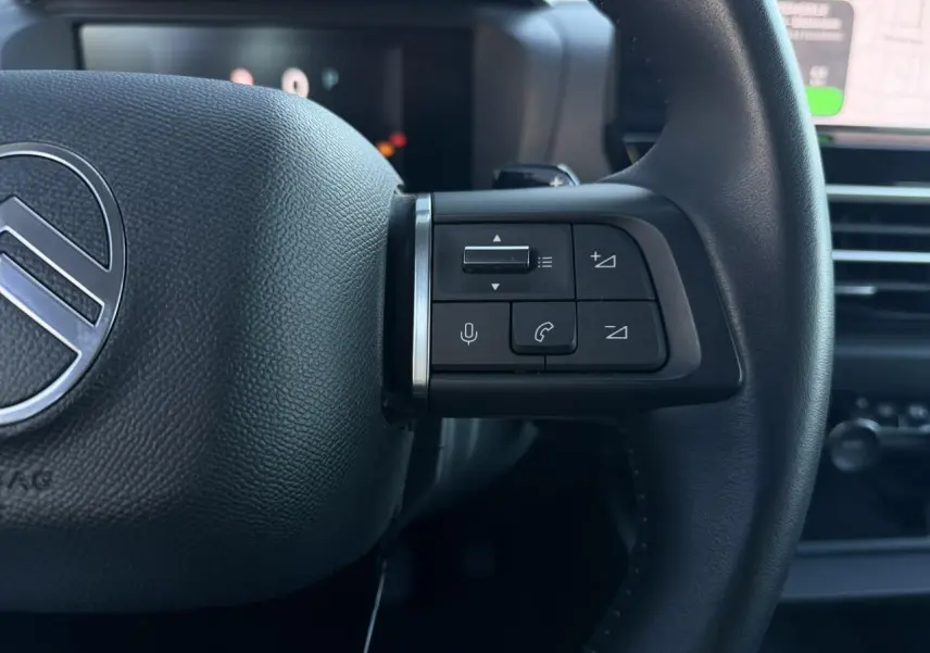 Gros plan sur les commandes du volant en cuir noir d'une Citroën C4 Hybrid 2025 avec écran tactile et navigation visible en arrière-plan.