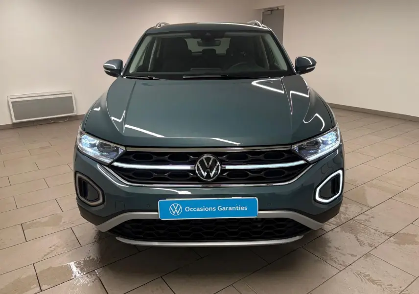 Vue frontale d'un Volkswagen T-Roc 1.5 TSI bleu pétrole avec éclairage LED et calandre chromée dans un showroom.