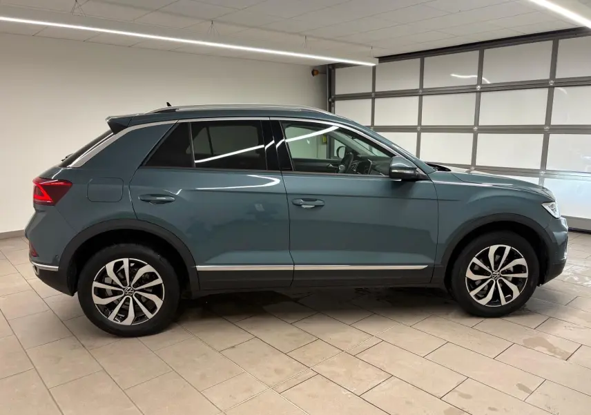 Vue de profil côté gauche d'un Volkswagen T-Roc bleu pétrole 2025 dans un garage, roues alliage visibles.