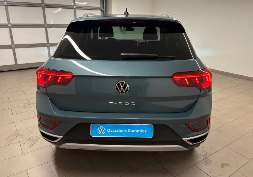 Vue arrière d'un Volkswagen T-Roc bleu pétrole 2025 dans un garage, avec feux arrière allumés et plaque "Occasions Garantis".