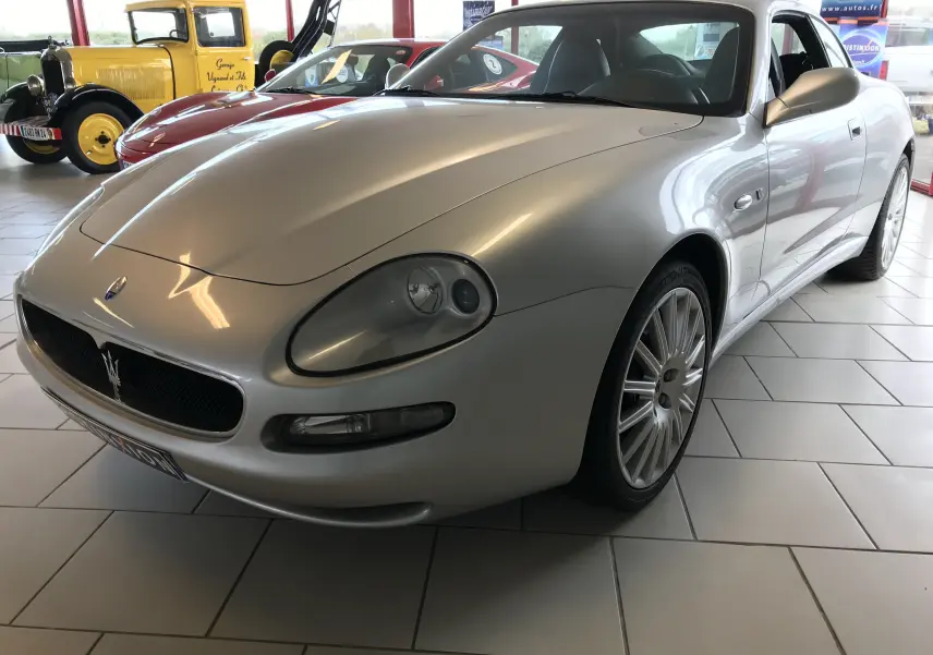 Maserati Coupé gris clair vue 3/4 avant droit, mettant en valeur ses phares ovales et la calandre au trident.