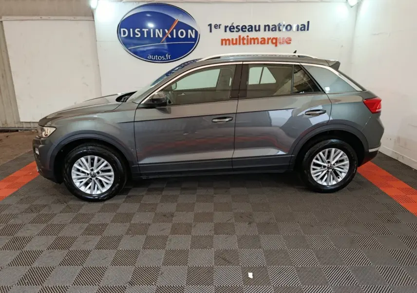 Profil côté gauche d'un Volkswagen T-Roc gris foncé 2021 dans un showroom avec sol damier et logo Distinxion en arrière-plan