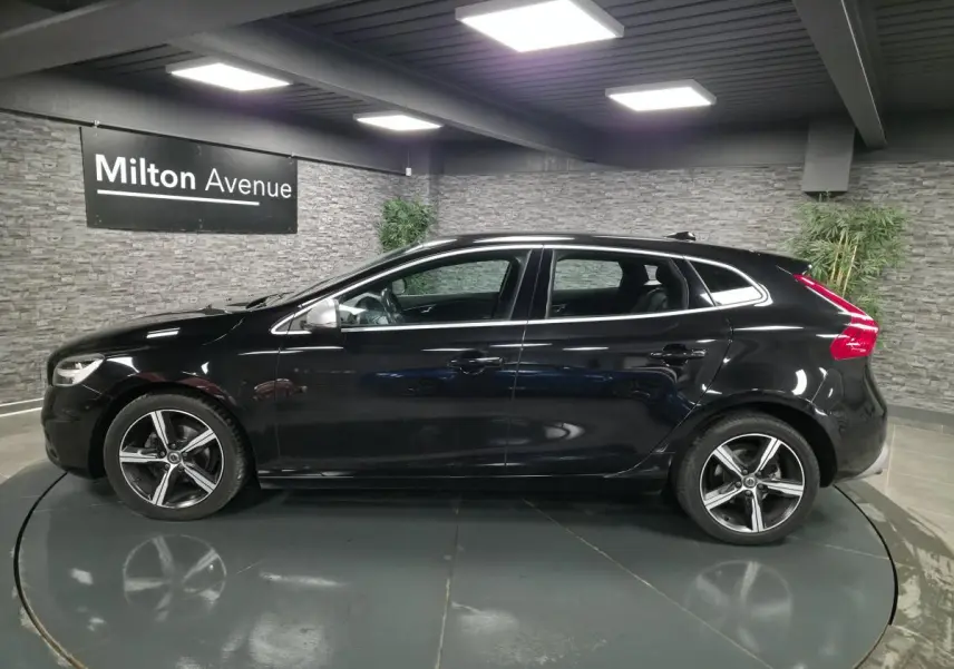 Profil côté gauche d'une Volvo V40 D2 R-Design noire métallisée avec jantes alliage Ixion 17 pouces dans un showroom.