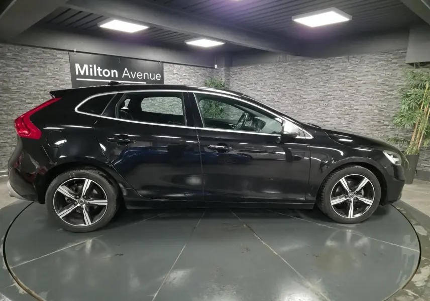 Profil côté gauche d'une Volvo V40 D2 R-Design noire avec jantes alliage Ixion 17 pouces dans un showroom.