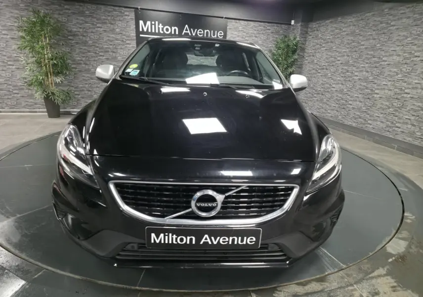 Vue avant d'une Volvo V40 D2 R-Design noire avec calandre chromée et plaque "Milton Avenue" en intérieur showroom.