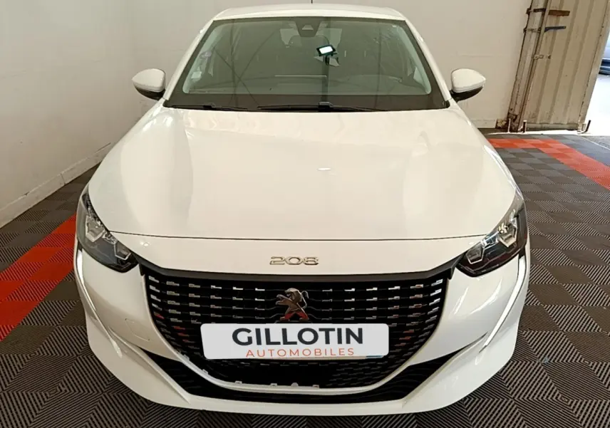Vue avant d'une Peugeot 208 blanche avec calandre noire et logo lion au centre, stationnée en intérieur.