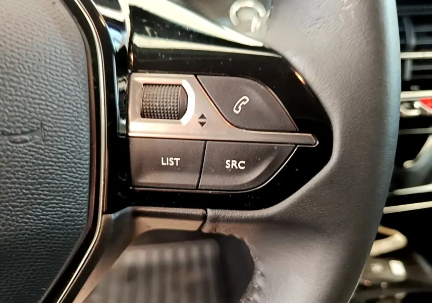 Gros plan sur les commandes gauche du volant cuir noir du Peugeot 208 blanc, avec boutons de gestion audio et appel.