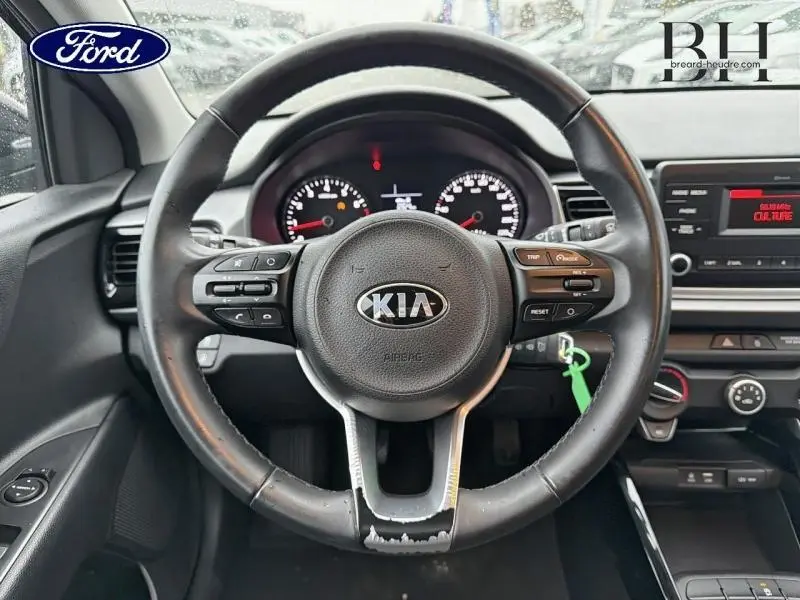 Vue intérieure centrée sur le volant cuir noir de la Kia Rio 2018 avec commandes audio et tableau de bord visible.