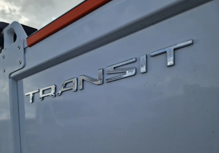 Gros plan sur l'arrière blanc d'un Ford Transit avec l'inscription "TRANSIT" en lettres chromées, sous un ciel nuageux.