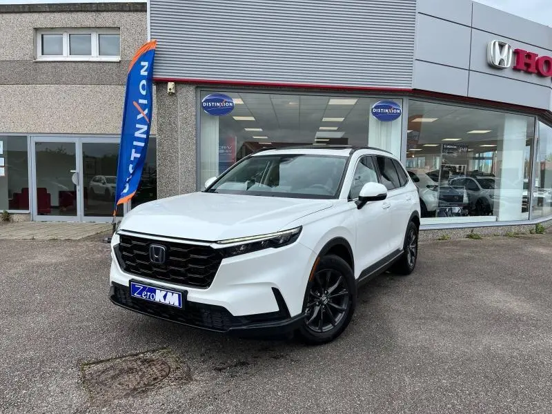 Honda CR-V blanc platine nacré vu en 3/4 avant droit devant un concessionnaire avec jantes noires et toit panoramique.