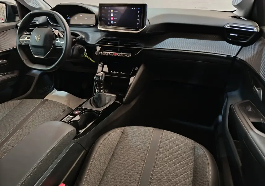 Vue intérieure avant droite du Peugeot 208 gris clair, mettant en valeur le tableau de bord digital et la console centrale avec boîte manuelle.