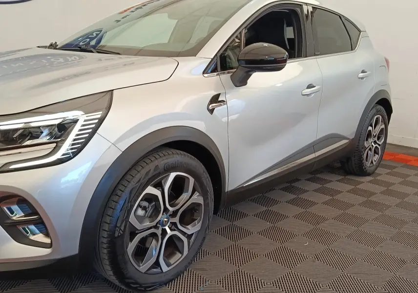 Vue 3/4 avant droit d'un Renault Captur Intens E-TECH gris clair avec jantes alliage et feux LED allumés.