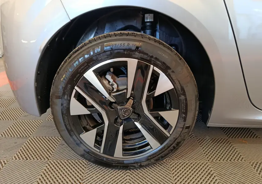 Gros plan sur la roue avant gauche de la Peugeot 208 gris clair, mettant en valeur la jante noire et argentée au design moderne.