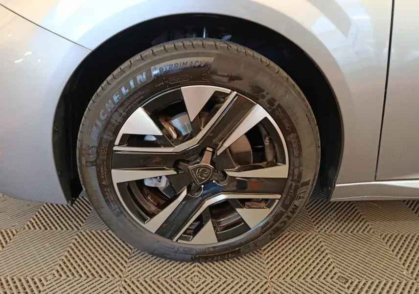 Gros plan sur la roue avant gauche d'une Peugeot 208 gris clair, mettant en valeur la jante alliage au design moderne.