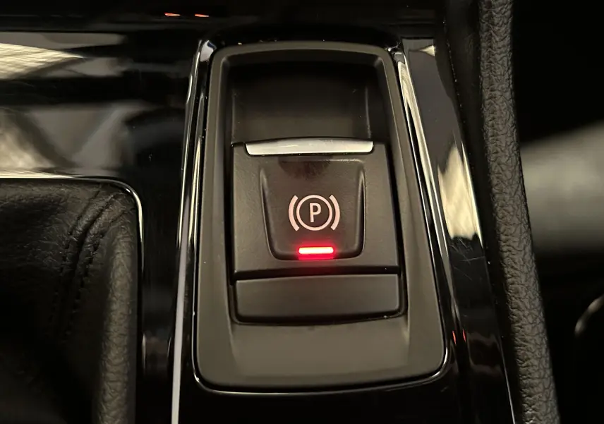 Gros plan sur le bouton de frein à main électronique éclairé en rouge dans la console centrale d'une BMW Série 2 Active Tourer blanche.