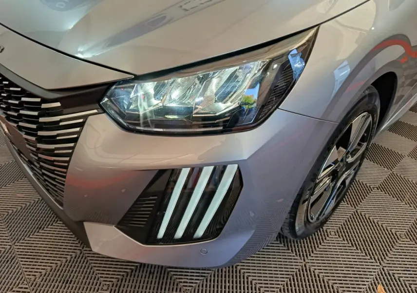 Gros plan sur l'avant droit gris clair du Peugeot 208 2025 avec phares LED et signature lumineuse verticale distinctive.