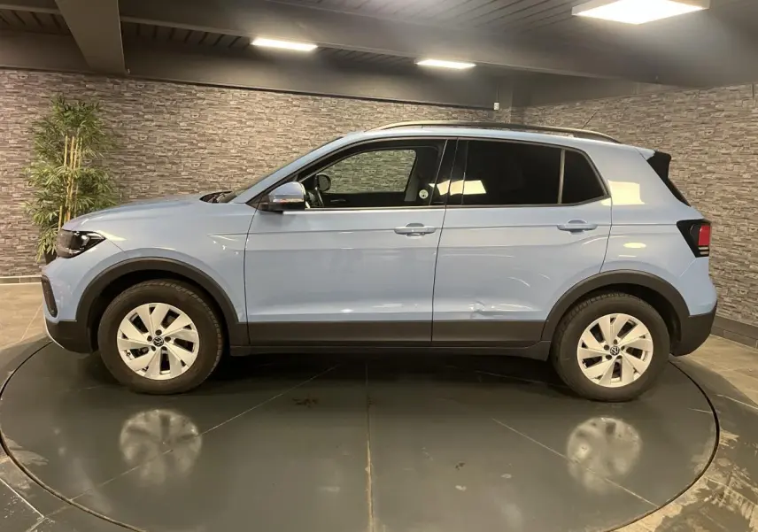 Profil côté gauche du Volkswagen T-Cross bleu clair 2024 avec vitres arrière surteintées en intérieur showroom.