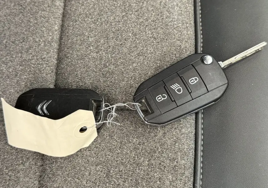 Clé de voiture noire Citroën posée sur siège tissu gris et cuir noir, avec porte-clés et étiquette blanche.