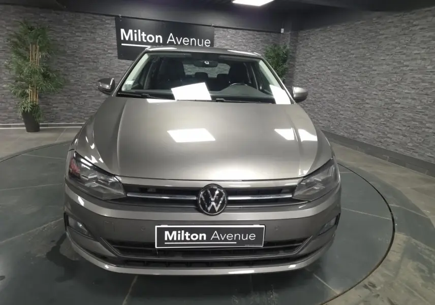 Volkswagen Polo gris métallisé vue de face en intérieur avec logo VW et plaque Milton Avenue visible.