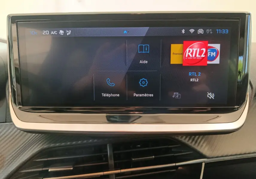 Écran tactile central affichant la radio RTL2 dans l'habitacle d'une Peugeot 208 gris foncé, vue frontale rapprochée.
