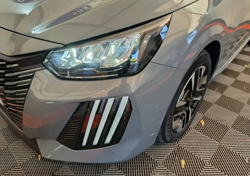 Gros plan sur l'avant droit gris foncé de la Peugeot 208 2025, mettant en valeur ses phares LED et jante noire.