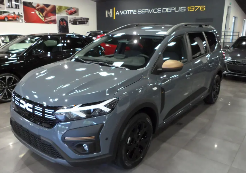 Vue 3/4 avant d'un Dacia Jogger TCe 110 gris urbain avec rétroviseurs beige et jantes noires en showroom.