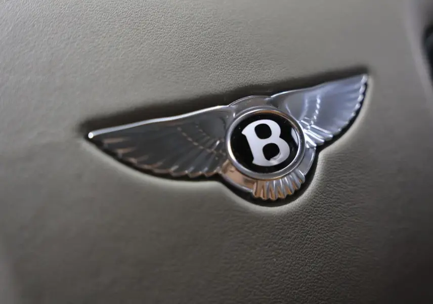Gros plan sur l'emblème Bentley argenté en forme d'ailes sur fond cuir beige, intérieur d'une Continental Flying Spur.