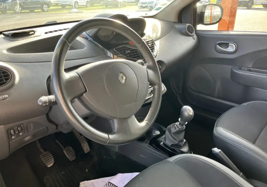 Vue intérieure côté conducteur d'une Renault Twingo II noire de 2011, avec volant, tableau de bord et levier de vitesses manuel visibles.