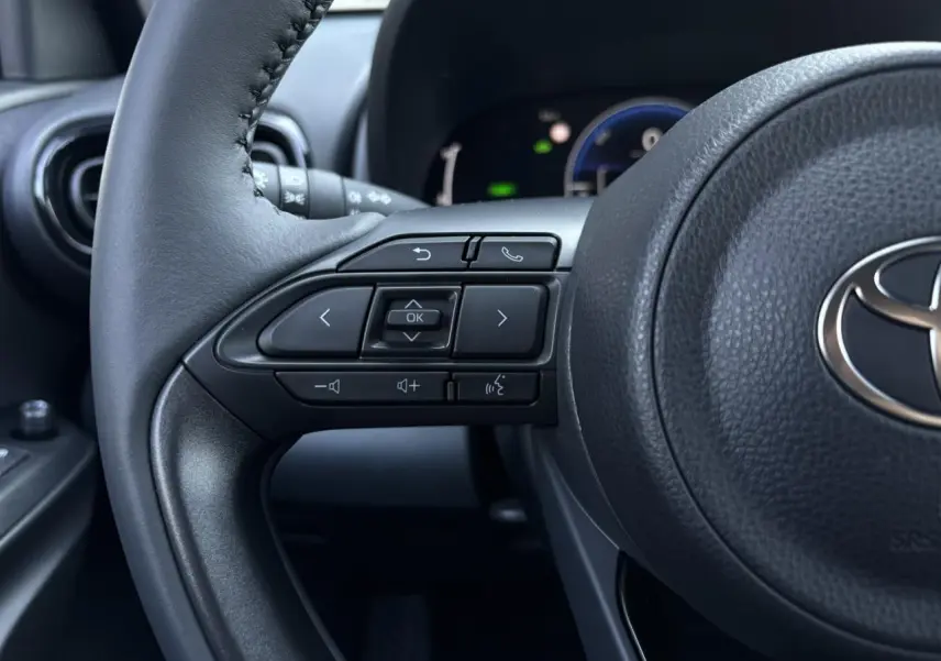 Gros plan sur le volant cuir noir de la Toyota Yaris 2025, avec commandes intégrées et tableau de bord numérique en arrière-plan.