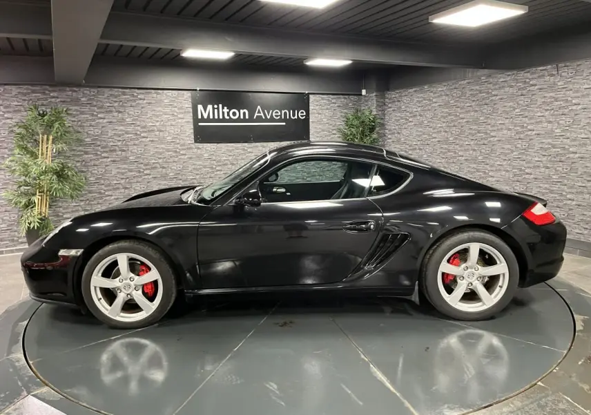 Profil côté gauche d'une Porsche Cayman noire 2008 avec jantes argentées et étriers de frein rouges en intérieur showroom.