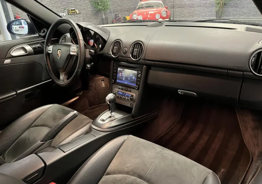 Intérieur noir de Porsche Cayman 987 2.7i 2008 vu côté conducteur avec console centrale et volant Porsche visible.