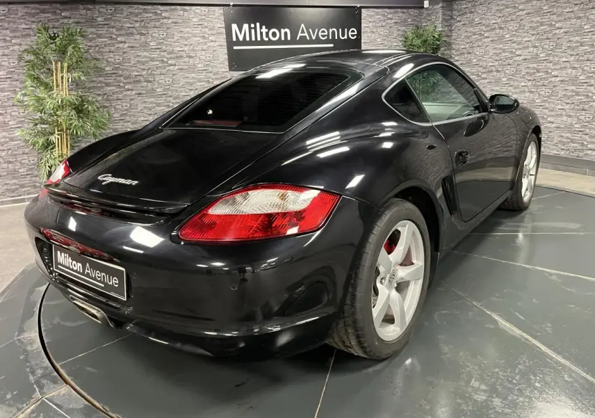 Vue 3/4 arrière droite d'une Porsche Cayman noire avec jantes 18 pouces et feux arrière rouges dans un showroom.