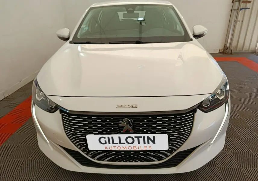 Vue avant d'une Peugeot 208 électrique blanche avec calandre noire et feux LED, en intérieur sur sol à damier rouge et gris.