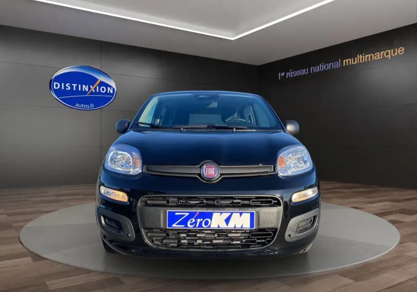 Vue frontale d'une FIAT Panda noire 2025 avec phares allumés et plaque "Zéro KM" en showroom.