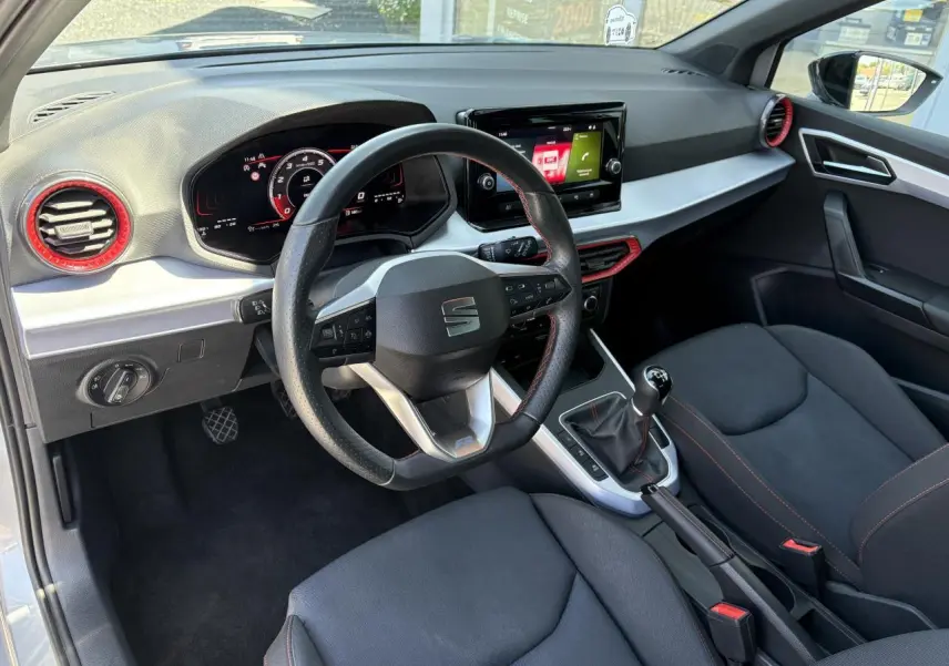 Vue intérieure côté conducteur du SEAT Arona 2024, volant sport cuir noir, inserts rouges autour des aérateurs et console centrale.
